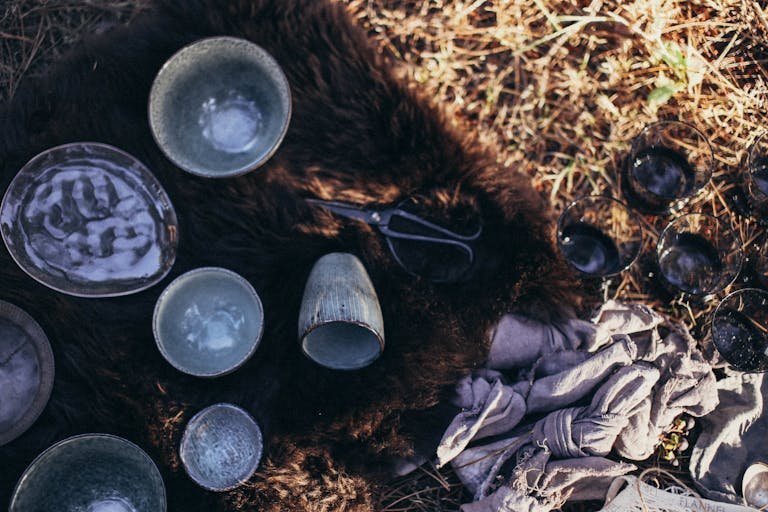 Overhead of composition with ceramic and glass utensil near scissors placed on fur on dry grass on sunny day