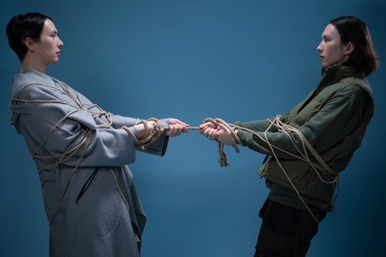 Two adults engaged in a symbolic tug of war, wrapped in rope against a blue background.